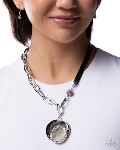 A close-up of a model wearing a silver-toned pendant necklace with a black ribbon strap, a small pink floral accent, and a round silver medallion pendant.