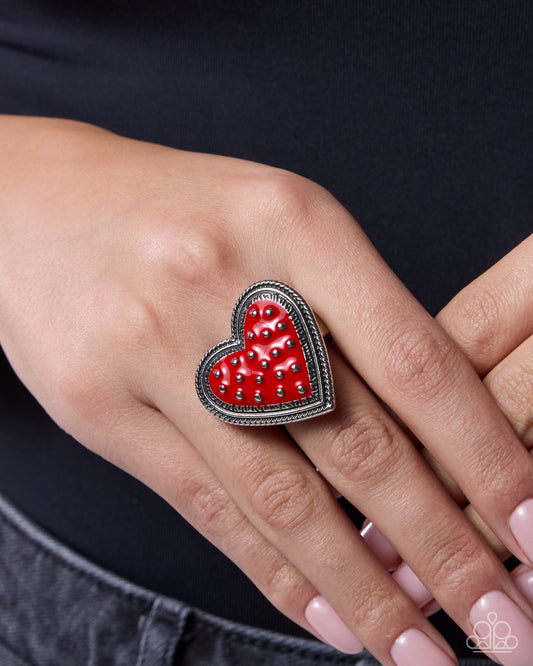 Red heart-shaped ring on a hand against a black background