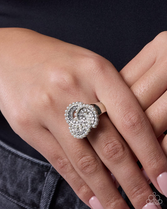 A close-up of a hand wearing a silver-toned ring with a large, swirling floral or spiral design set with clear stones. The hand is resting against a dark navy shirt and gray denim.