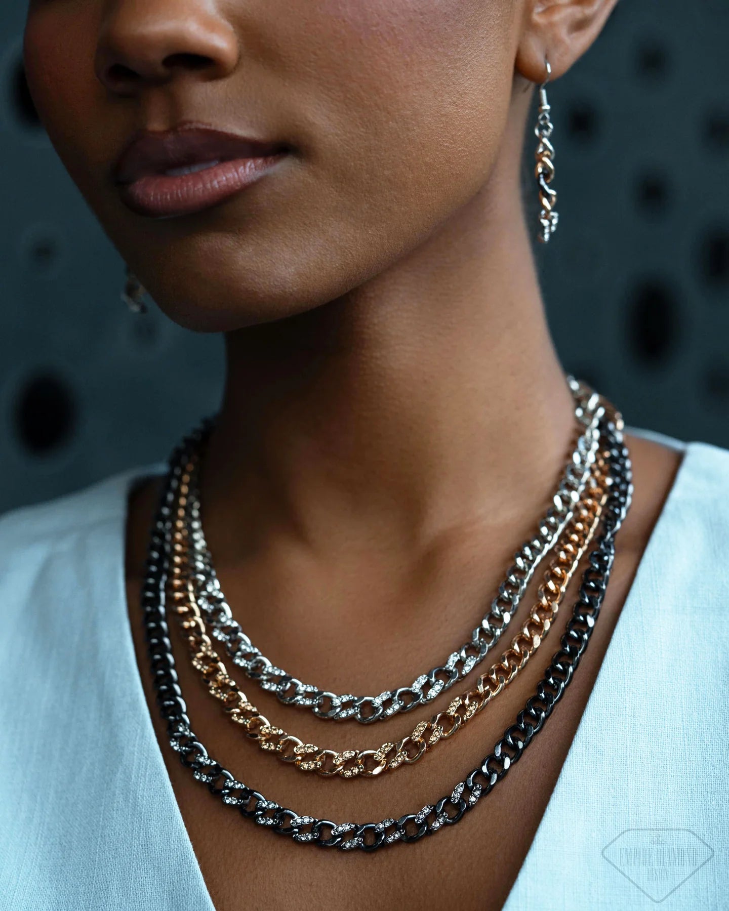 Close-up of a model wearing four layered chain necklaces in silver, black, and gold tones, along with a matching dangling earring.