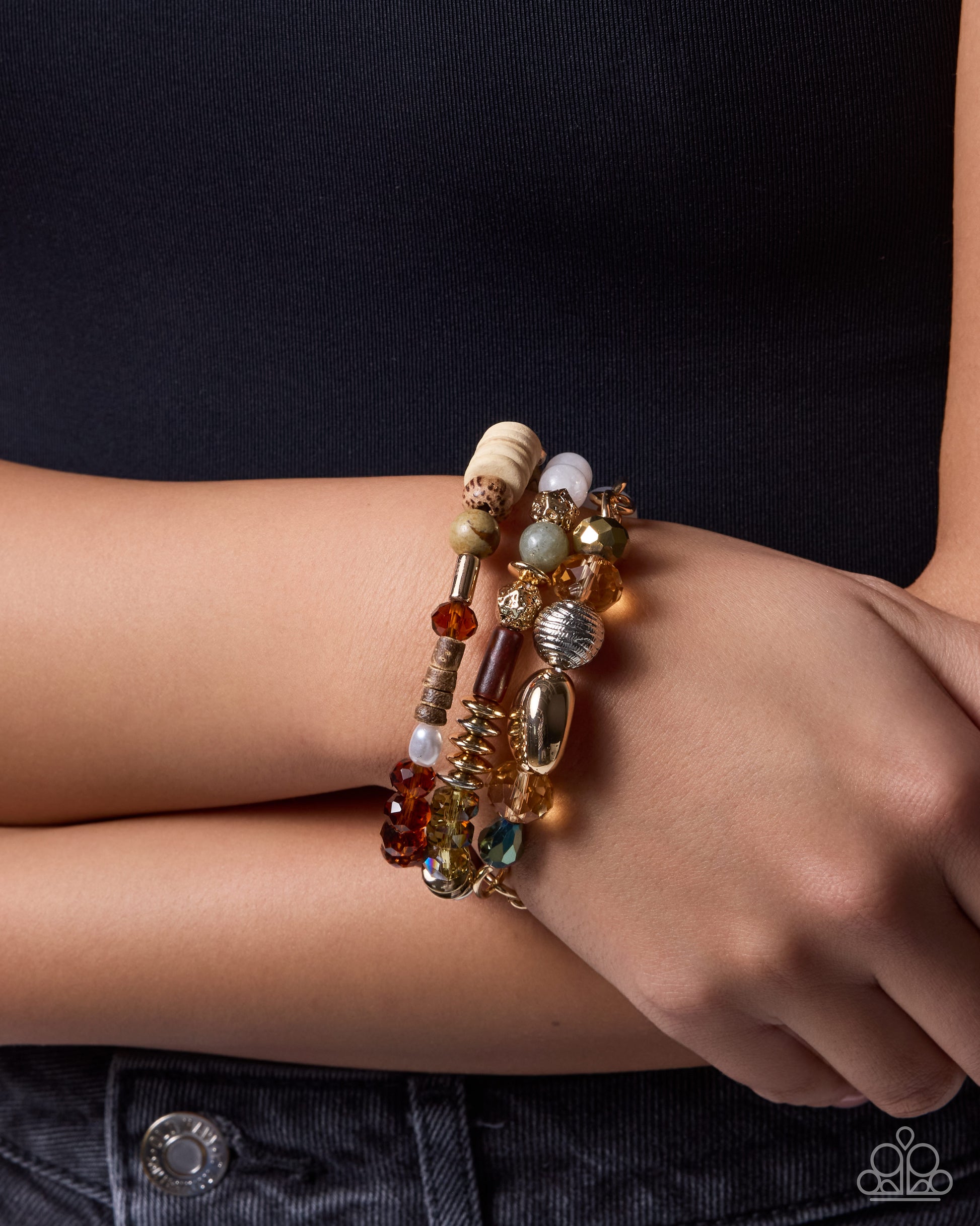 A close-up of a person's wrist wearing a stack of three beaded bracelets. The bracelets feature a mix of gold, brown, beige, and white beads with some translucent and metallic details, worn over a black top and dark denim jeans.