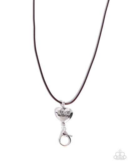 A silver heart-shaped pendant engraved with 'TRUST in Jesus' hanging from a dark brown cord necklace on a white background.