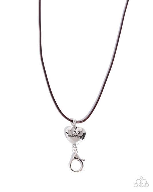 A silver heart-shaped pendant engraved with 'TRUST in Jesus' hanging from a dark brown cord necklace on a white background.