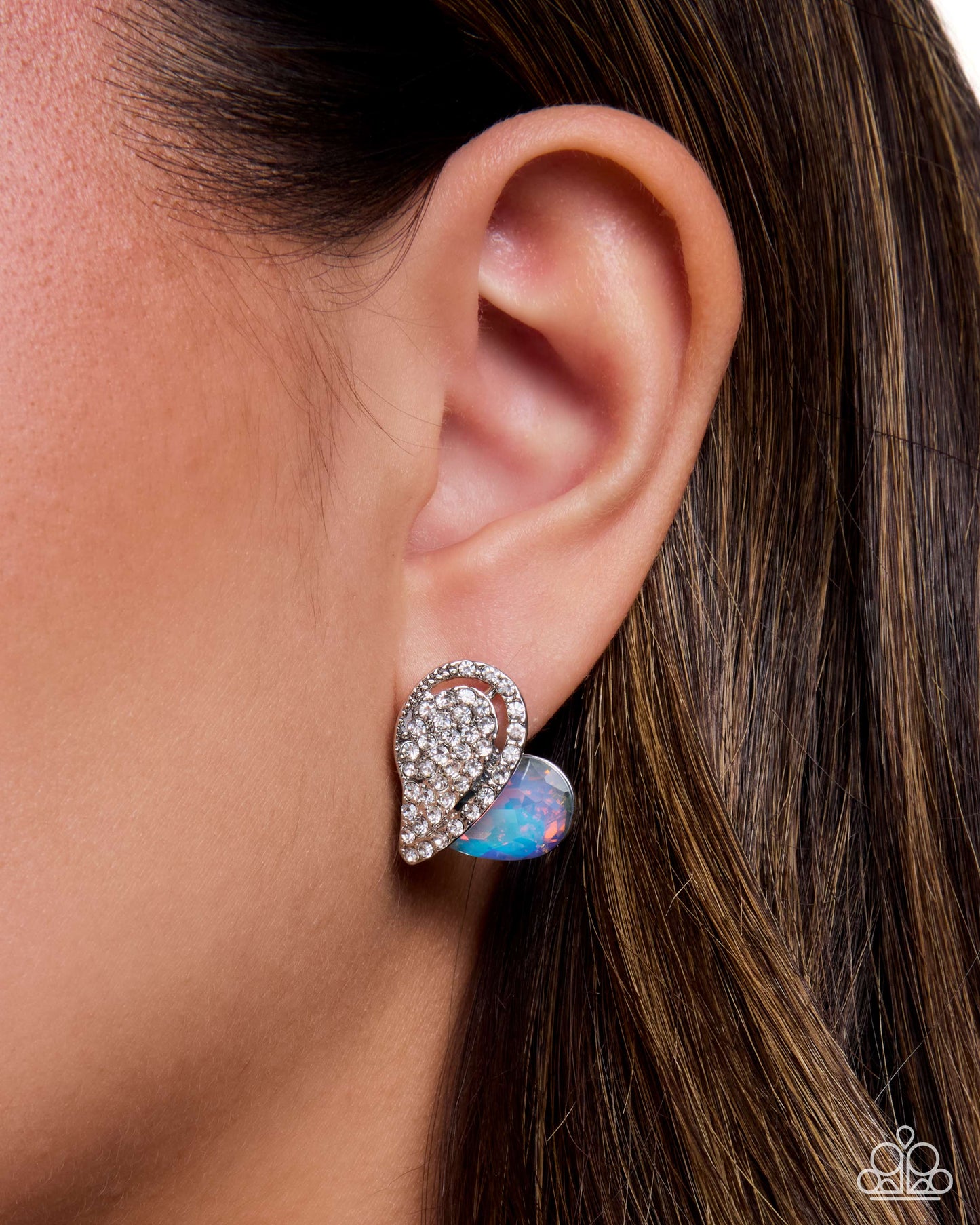 A close-up of a woman's ear wearing a silver-toned wing-shaped stud earring adorned with clear crystals and a small iridescent opal-like accent, against dark brown hair.