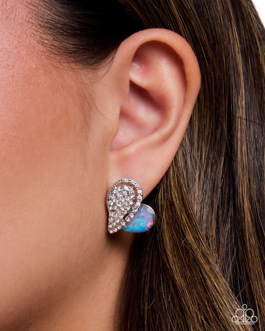 A close-up of a woman's ear wearing a silver-toned wing-shaped stud earring adorned with clear crystals and a small iridescent opal-like accent, against dark brown hair.