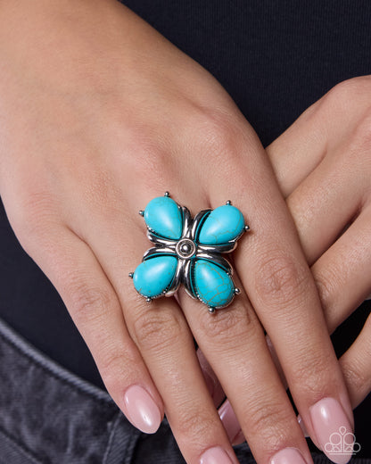 Turquoise flower-shaped ring on a person's finger against a neutral background
