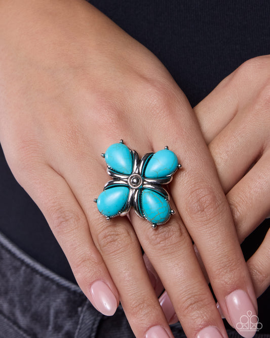 Turquoise flower-shaped ring on a person's finger against a neutral background