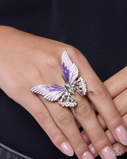 A silver butterfly ring with white and purple enamel wings and clear rhinestone accents, worn on a person's ring finger.