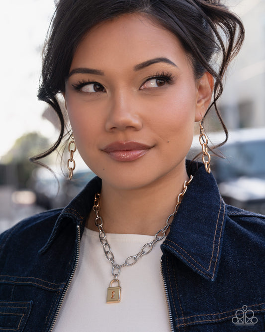 A close-up portrait of a woman wearing a silver chain necklace with a gold padlock pendant, matching gold drop earrings, and a dark denim jacket over a white top.