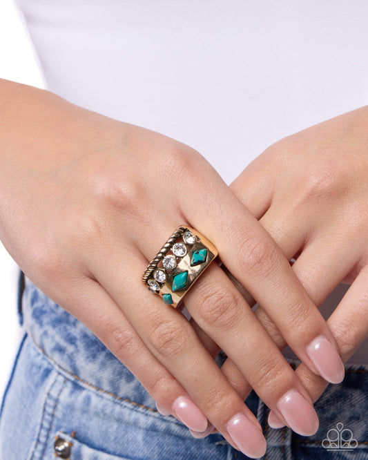 A close-up of a person's hand wearing a wide gold band ring with turquoise-colored stones and clear rhinestones, paired with a white top and blue jeans.