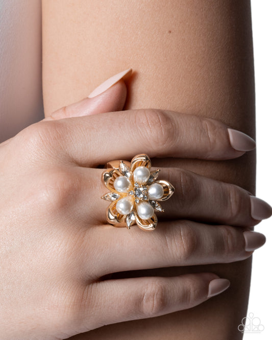A gold flower-shaped ring with white faux pearls and small clear crystals, worn on a woman's finger against a clean white background.