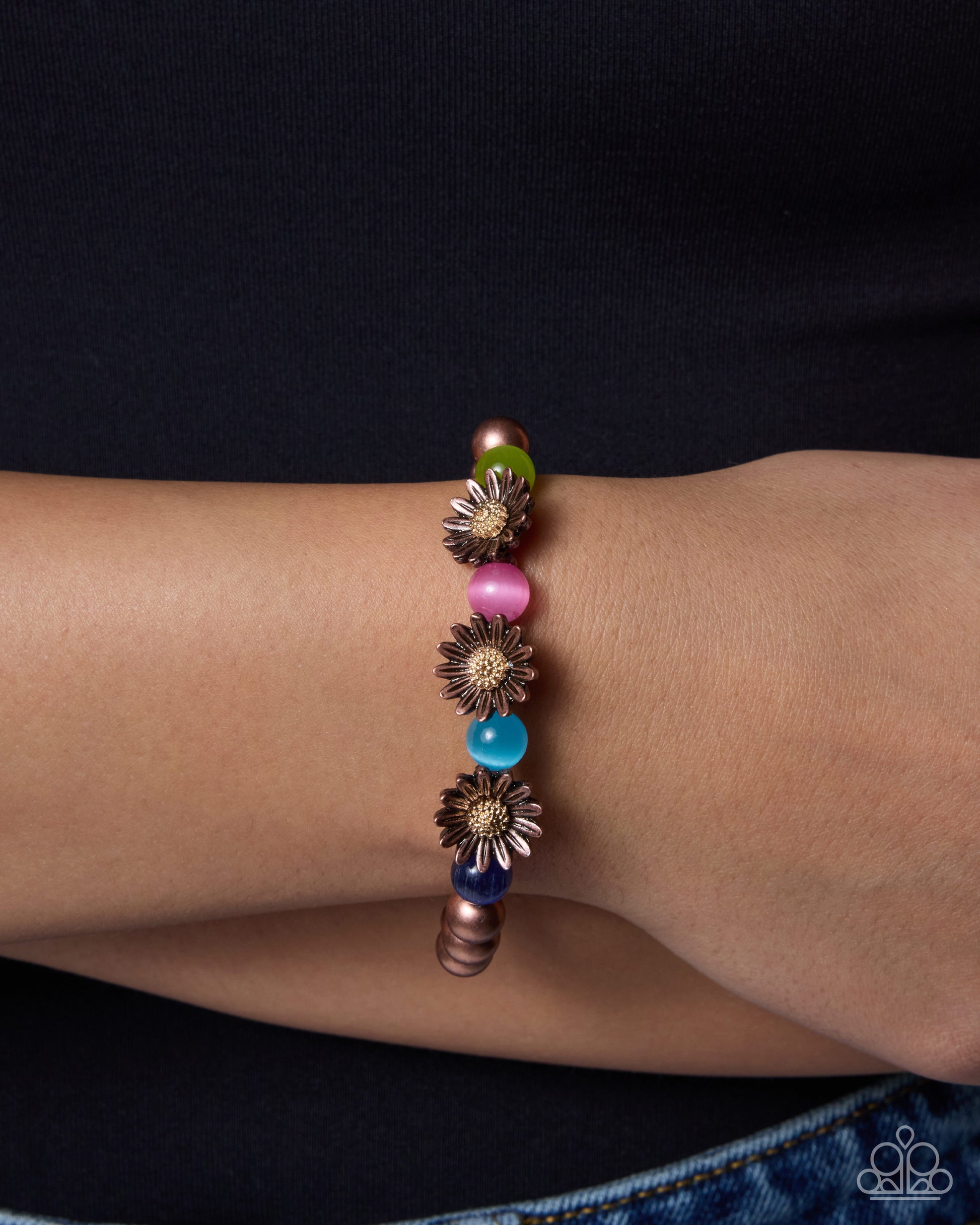 A close-up of a person's wrist wearing a beaded bracelet with metallic rose gold flower charms, pink and blue beads, and a green bead, set against a dark navy shirt.