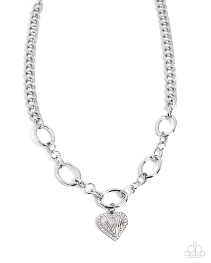 A silver-toned chain necklace featuring alternating oval and link-style segments, with a small heart-shaped charm at the center adorned with clear rhinestones. The necklace is displayed on a clean white background with a small Paparazzi logo watermark in the bottom right corner.