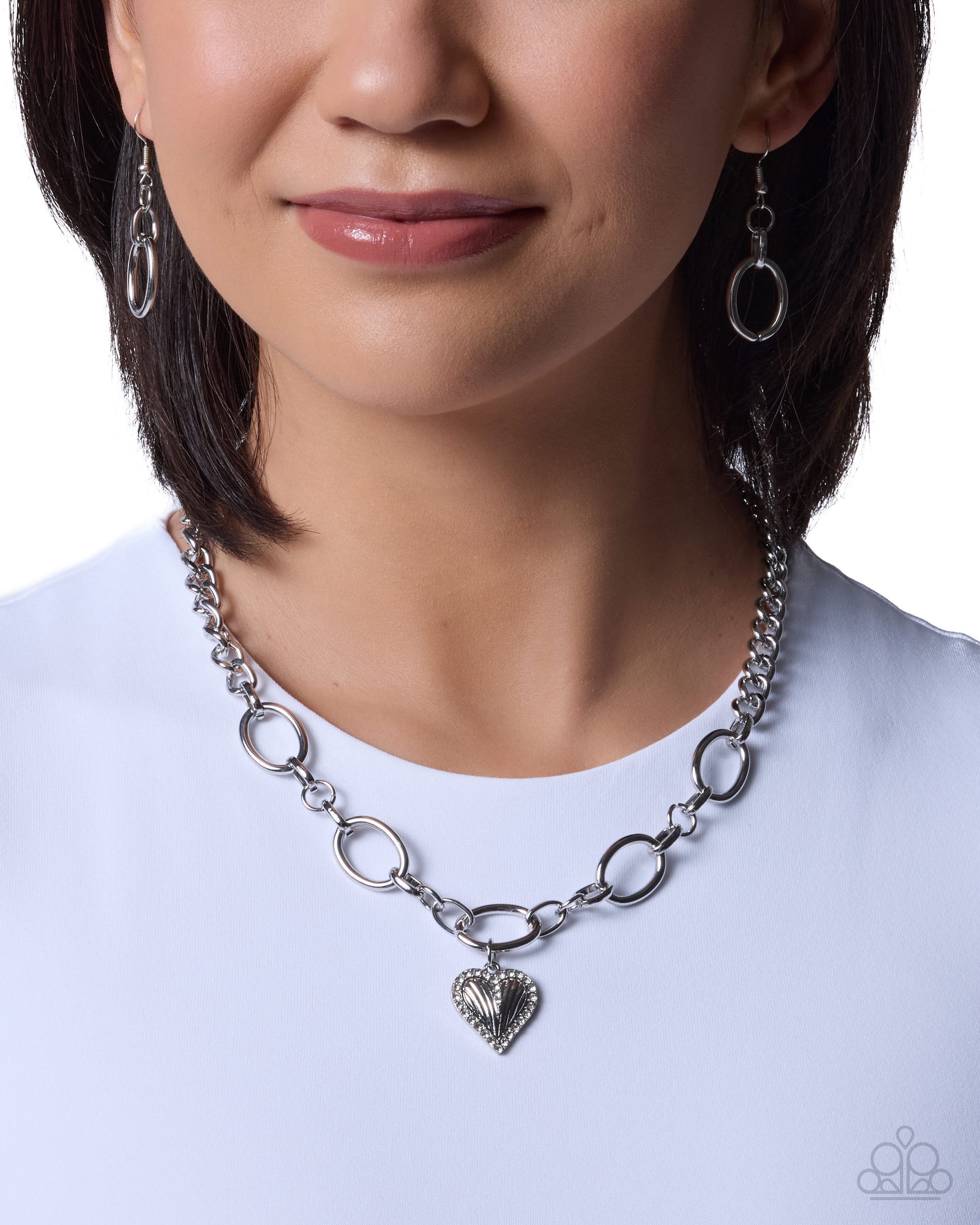 A close-up of a woman wearing a silver chain necklace with alternating oval and elongated links, featuring a textured heart pendant at the center. She is wearing a white top against a clean white background.