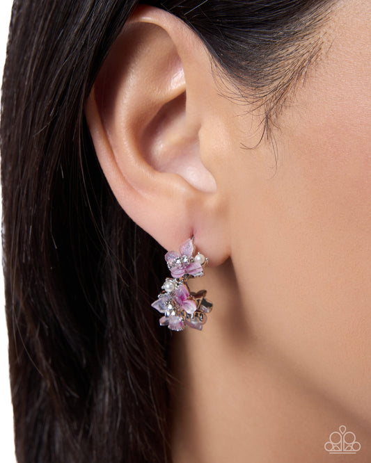 A close-up of a model's ear wearing a dangling floral earring with pink and lavender crystals and silver-toned metal.
