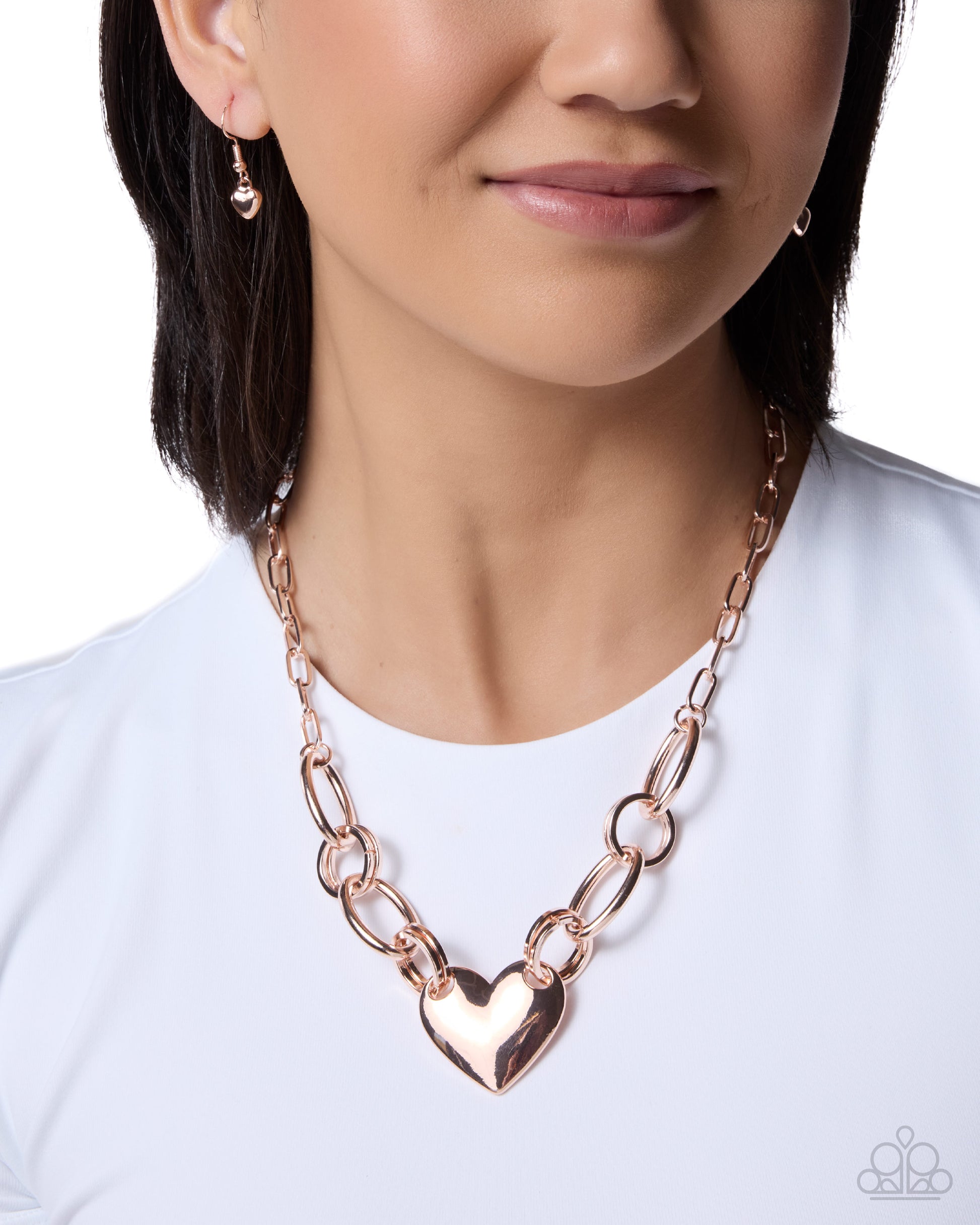 A close-up of a woman wearing a rose gold chain necklace with a heart-shaped pendant, styled with matching rose gold drop earrings against a white background.