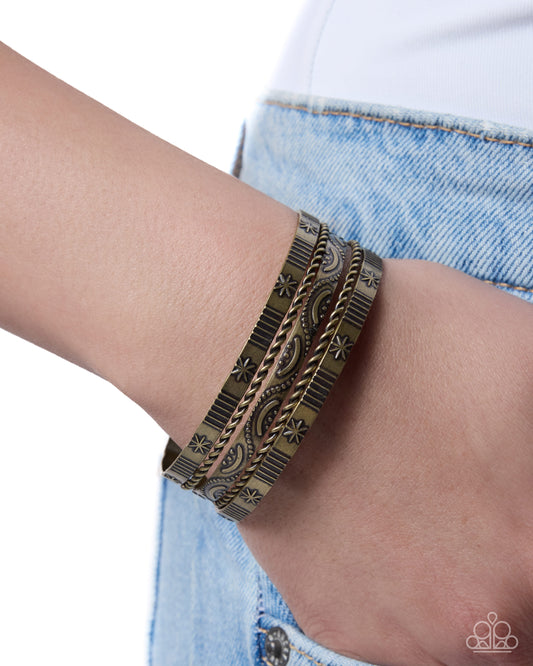 A close-up of a person's wrist wearing three stacked metallic cuffs with a distressed bronze and gold finish, paired with light blue denim jeans.