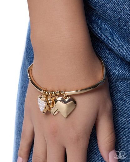 A gold bangle bracelet with three dangling heart charms, including one textured white heart and two polished gold hearts, shown worn on a person's wrist against a blue denim background.