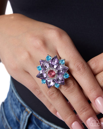 A close-up of a hand wearing a large flower-shaped ring with lavender and light blue gemstones surrounding a pink center, worn over a black top and blue jeans.