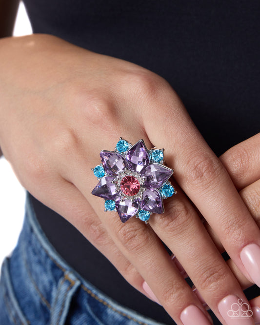 A close-up of a hand wearing a large flower-shaped ring with lavender and light blue gemstones surrounding a pink center, worn over a black top and blue jeans.