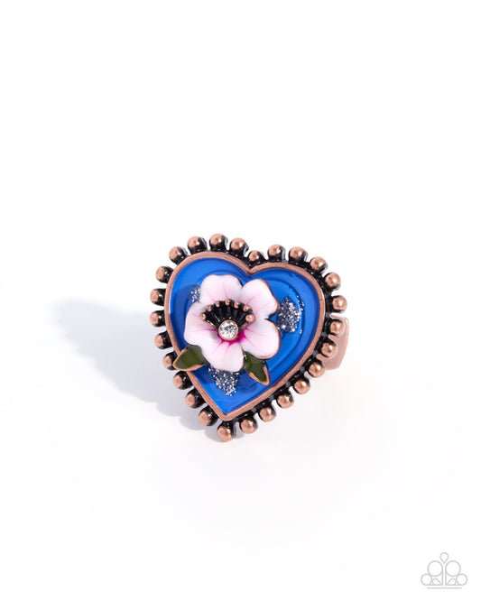 A heart-shaped ring featuring a vibrant blue enamel background, a central white and pink flower with a black center, and a copper-toned beaded border, shown on a white background.