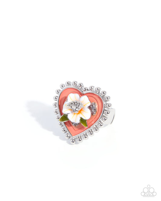 A heart-shaped silver ring featuring a coral pink enamel center, a white flower with green leaves, and a border of small clear rhinestones, shown on a white background.