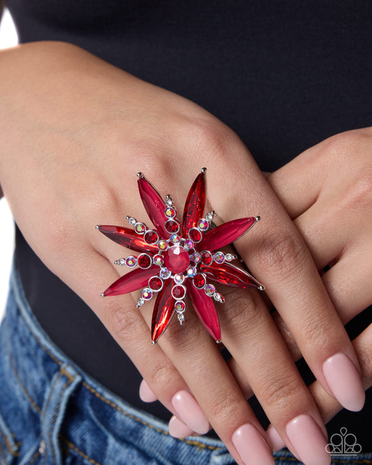 A close-up of a hand wearing a large, red flower-shaped ring with clear rhinestones, worn over a black top and blue jeans.
