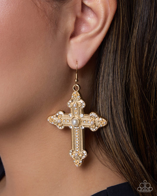 A close-up of a model's ear wearing a gold cross dangle earring embellished with clear rhinestones and a central pearl, with her dark brown hair visible.