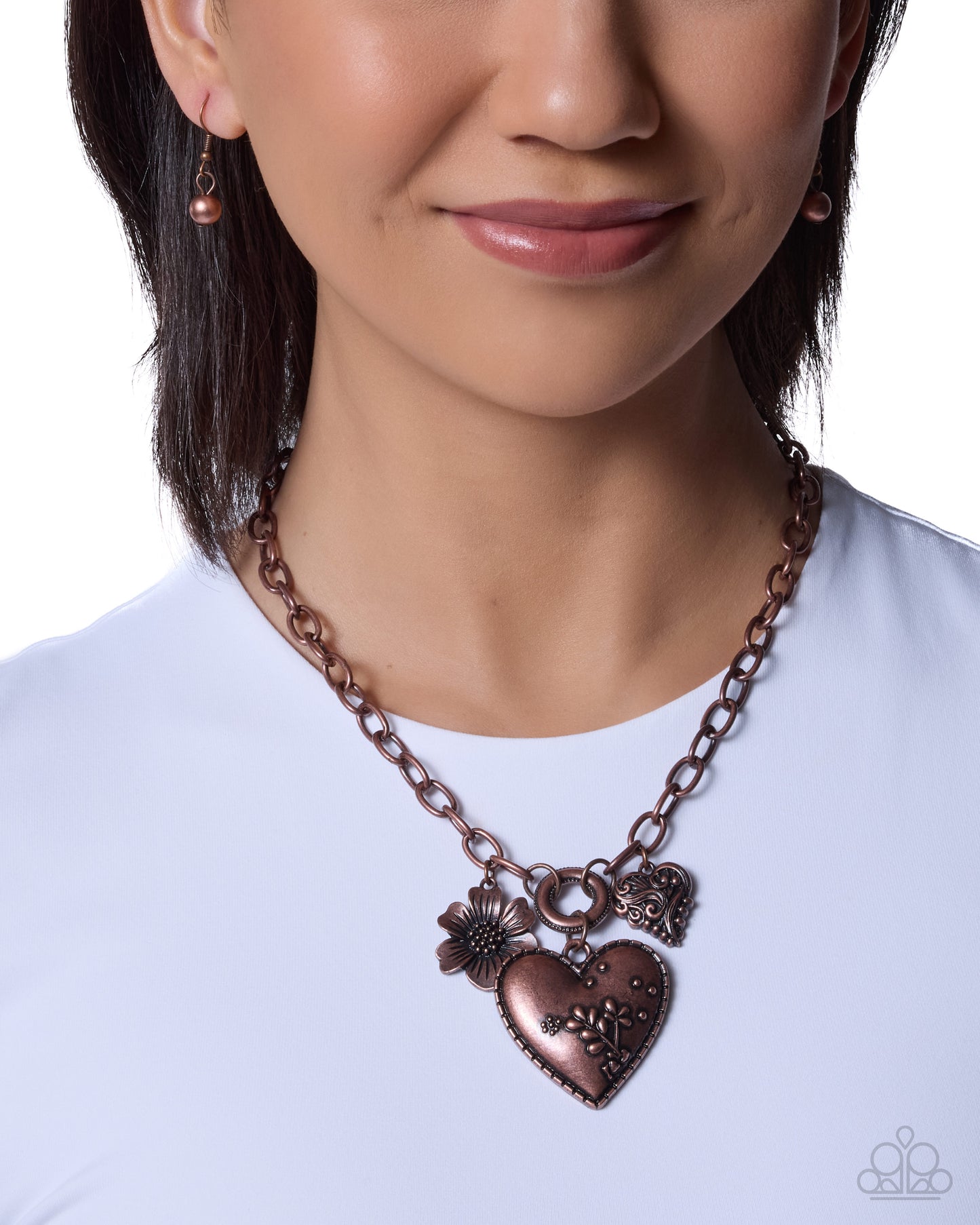 A close-up of a woman wearing a bronze-toned heart pendant necklace with a flower charm and small heart charms, paired with matching earrings against a white background.