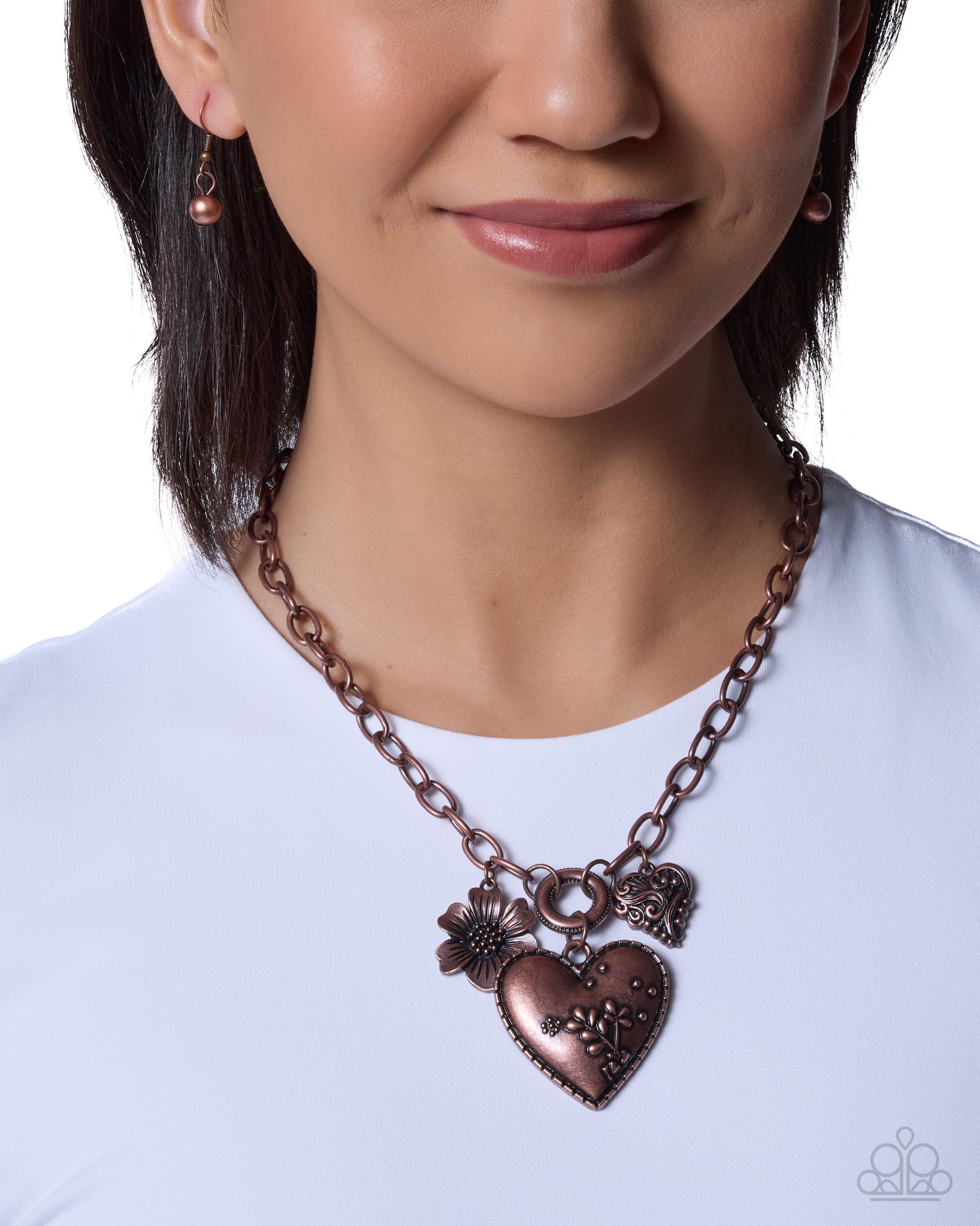 A close-up of a woman wearing a bronze-toned heart pendant necklace with a flower charm and small heart charms, paired with matching earrings against a white background.