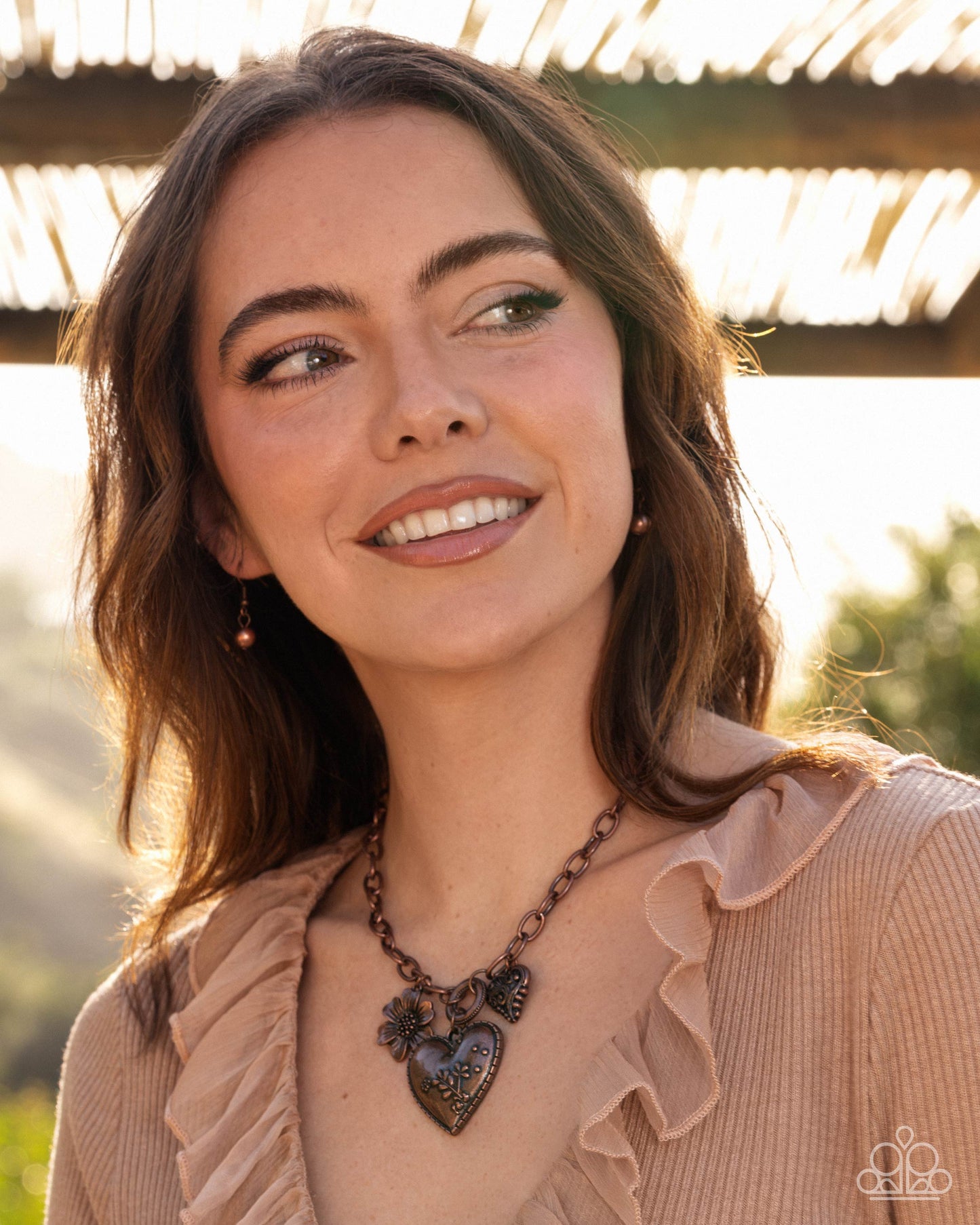 A woman with brown hair smiles while wearing a bronze heart pendant necklace with a floral design, styled with a beige ruffled top in an outdoor setting.