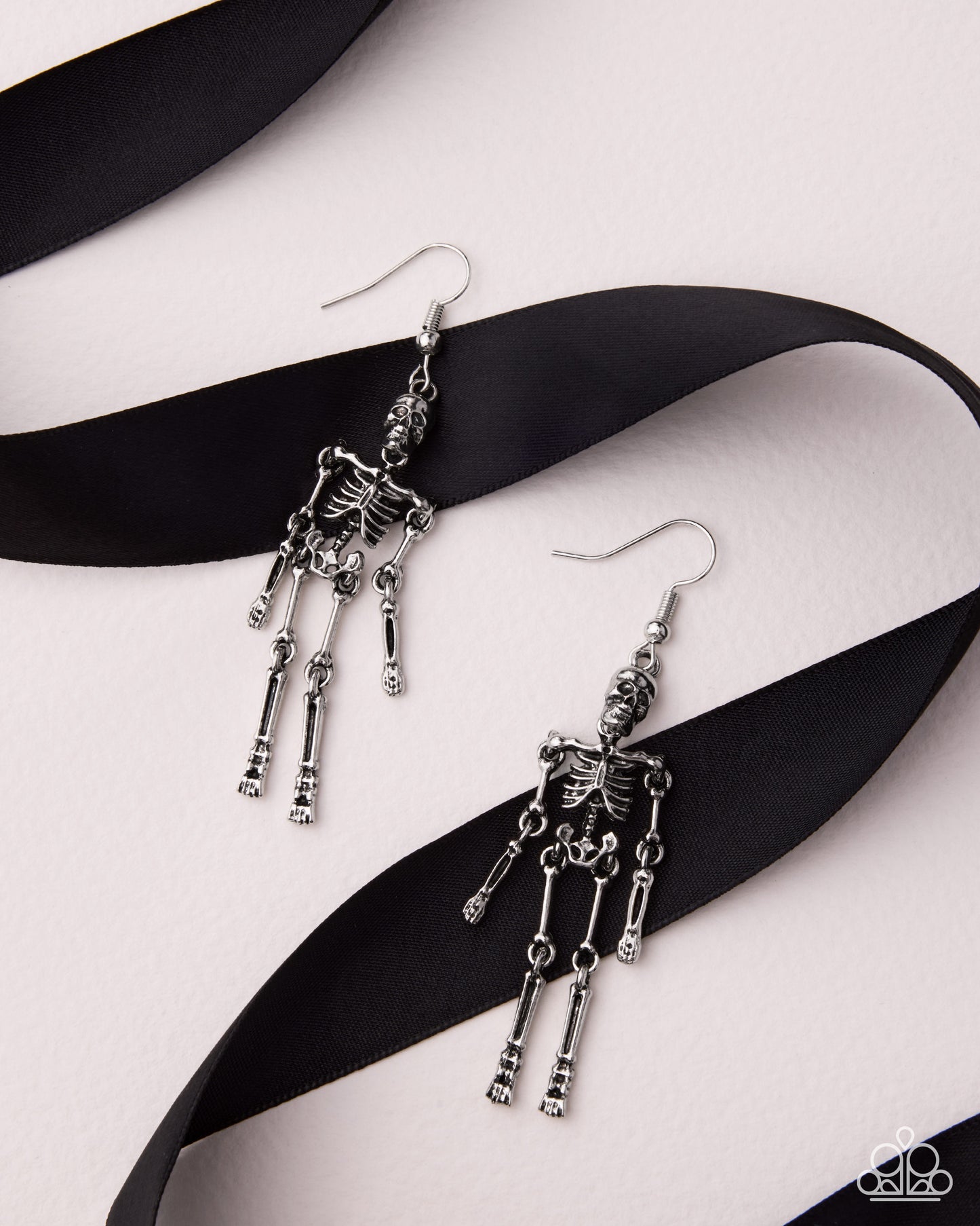 A pair of dangling skeleton earrings in silver-tone metal, displayed on a black ribbon over a light pink background.