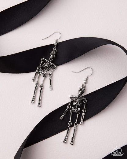 A pair of dangling skeleton earrings in silver-tone metal, displayed on a black ribbon over a light pink background.