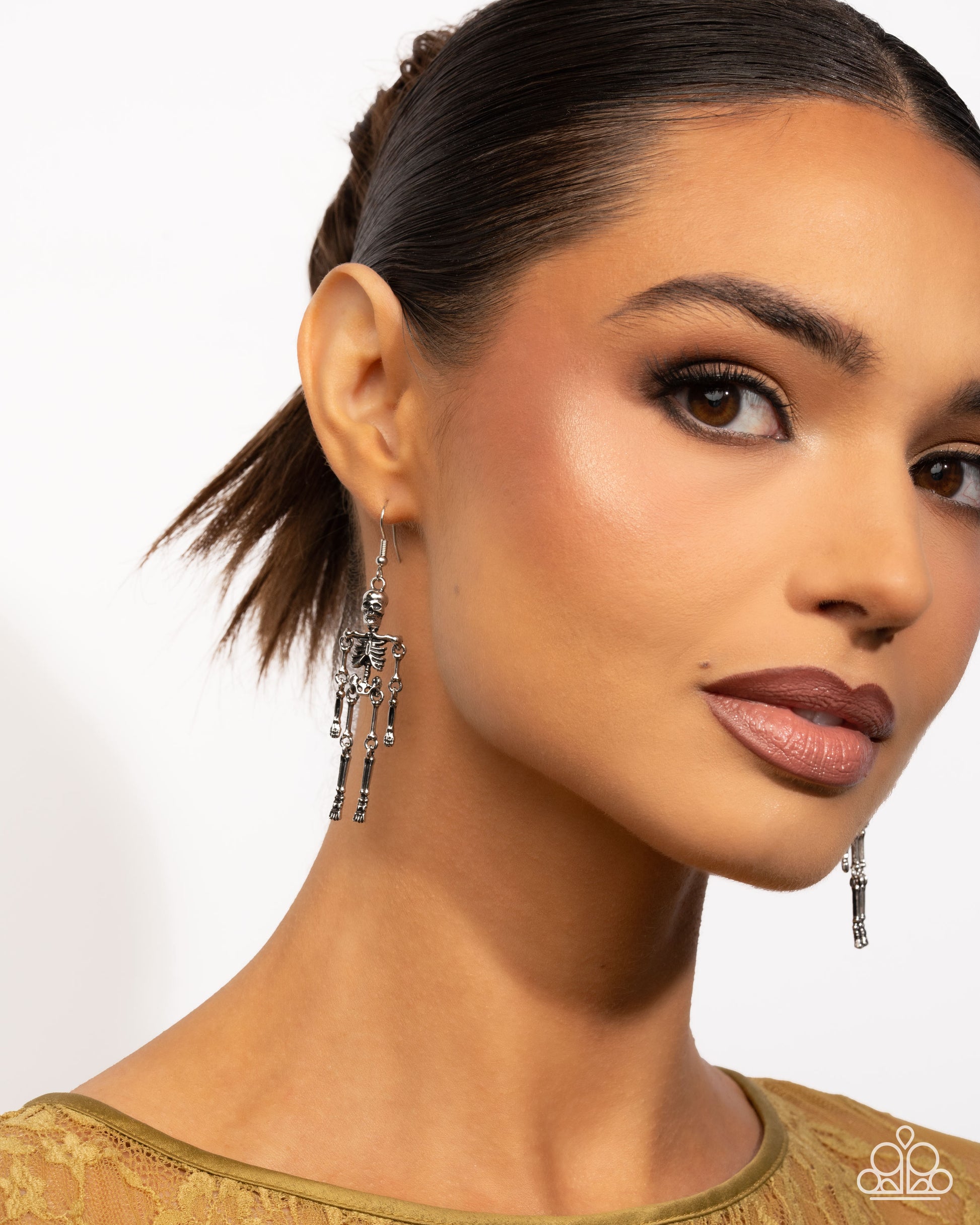 A close-up of a woman wearing long, dangling silver skeleton earrings, with her hair pulled back and a gold top visible against a clean white background.