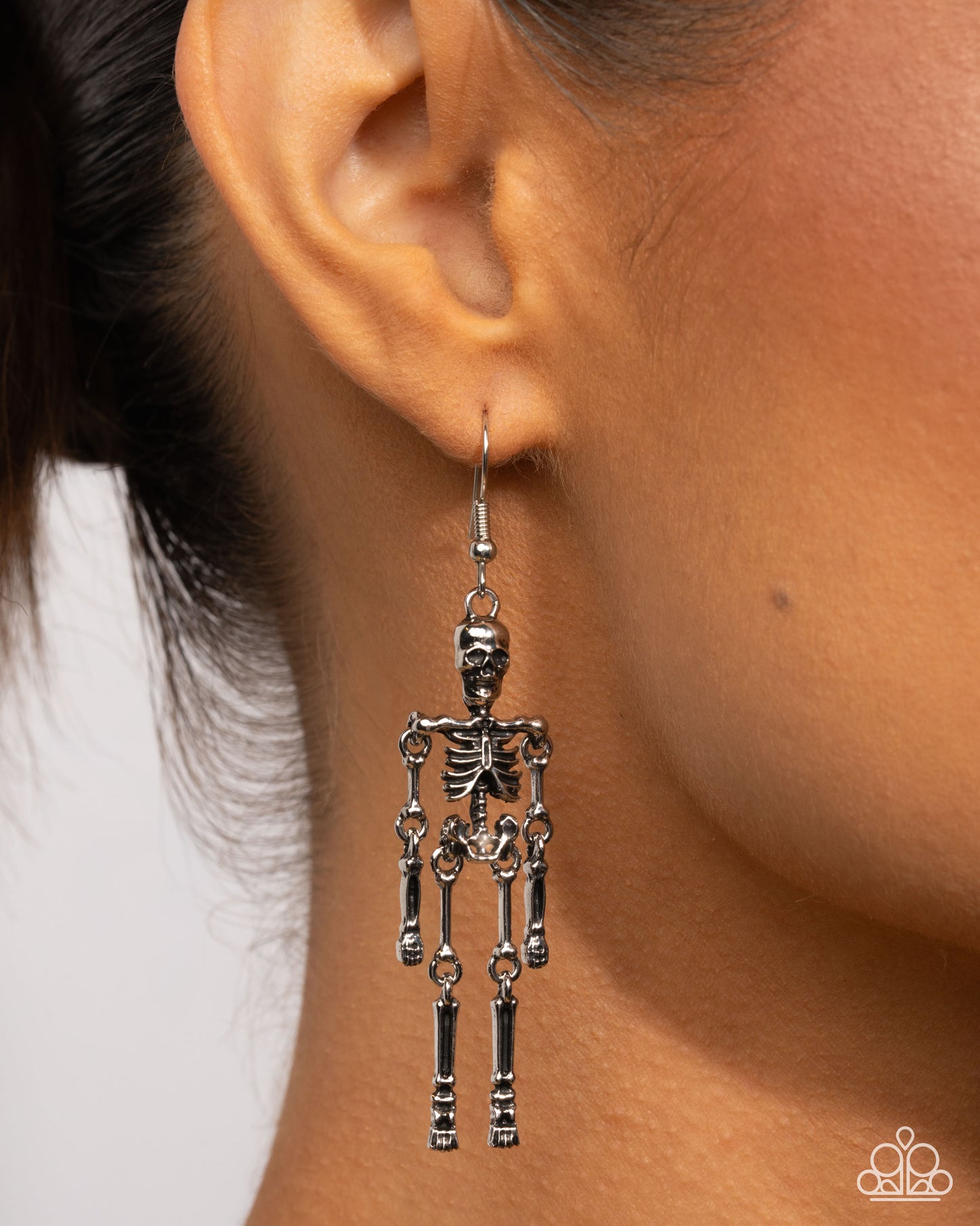A close-up of a model's ear wearing a dangling skeleton-shaped earring. The earring is made of silver-toned metal with articulated joint details.