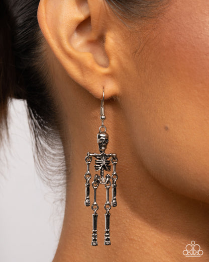 A close-up of a model's ear wearing a dangling skeleton-shaped earring. The earring is made of silver-toned metal with articulated joint details.
