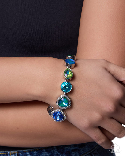 A close-up of a person's wrist and hand wearing a silver cuff bracelet with five colorful teardrop and oval gemstones in shades of blue, green, and dark blue.