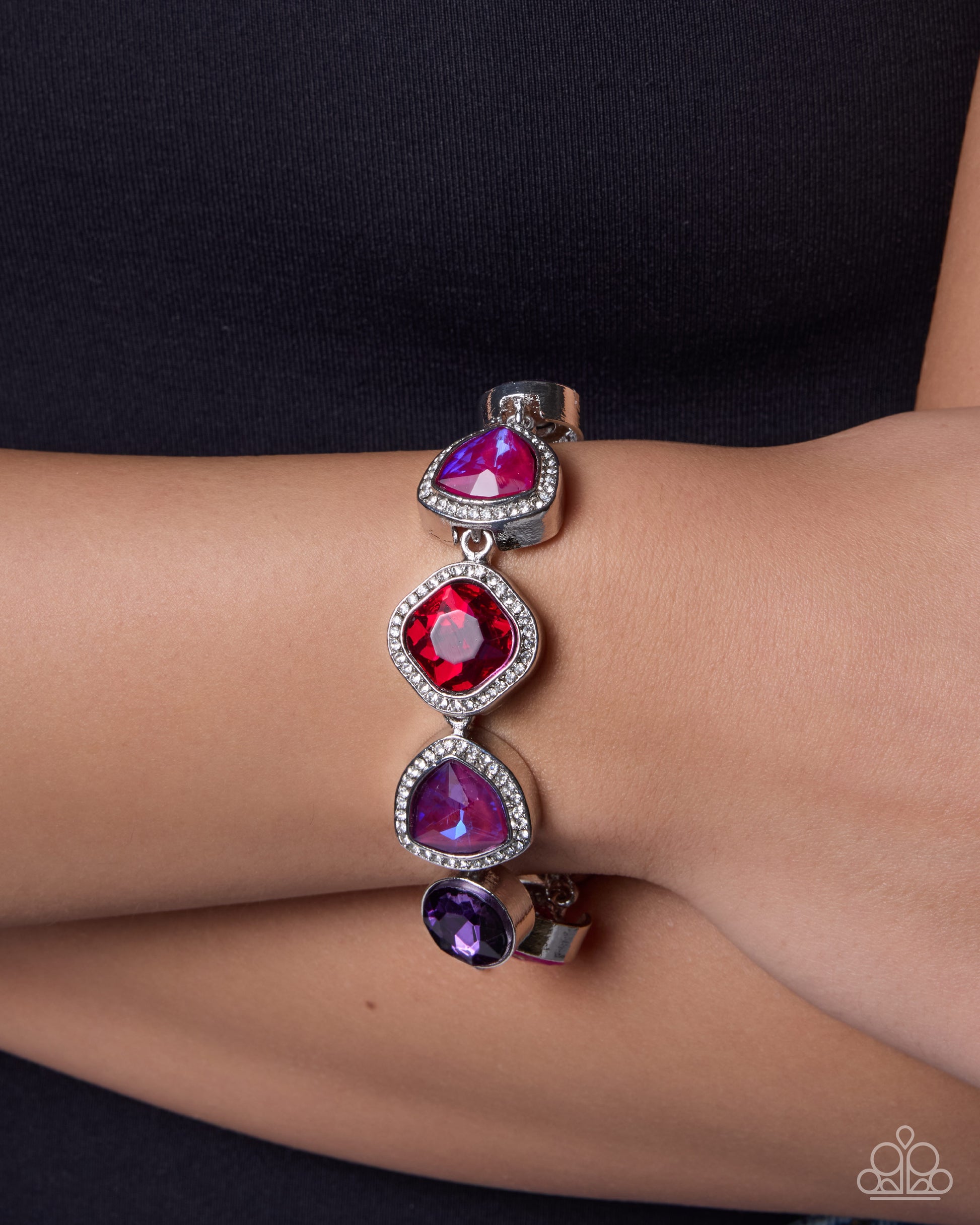 A close-up of a silver cuff bracelet with three vertical dangling gemstone charms in pink, red, and purple, worn on a person's wrist.