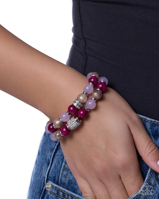 A close-up of a person's wrist wearing two beaded bracelets. The bracelets feature glossy magenta beads, pale lavender beads, and metallic champagne beads with a silver rhinestone-embellished spacer.