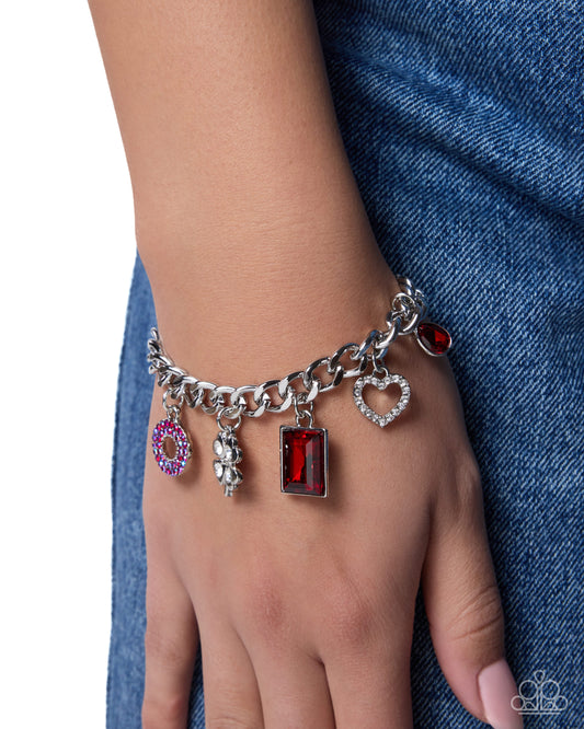 A silver charm bracelet with multiple dangling charms, including a heart, ring, and rectangular red gemstone charms, shown worn on a person's wrist against a blue denim background.