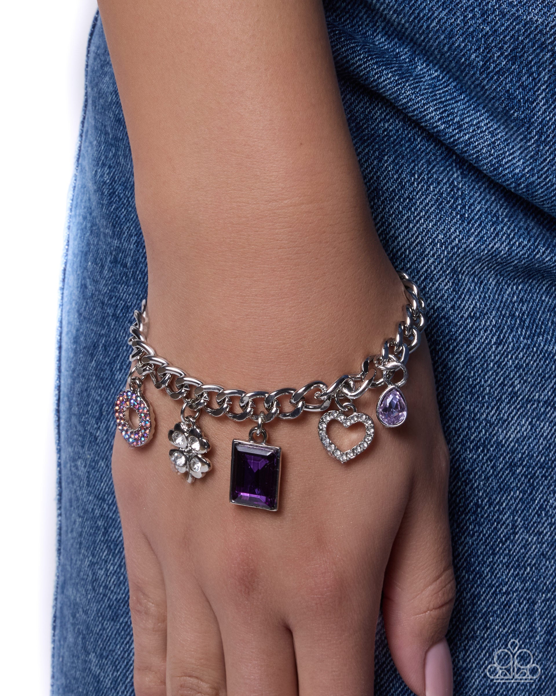 A silver-tone charm bracelet with multiple dangling charms, including a purple rectangular stone, a heart-shaped crystal, a flower, and a circular crystal charm, shown worn on a person's wrist against blue denim fabric.