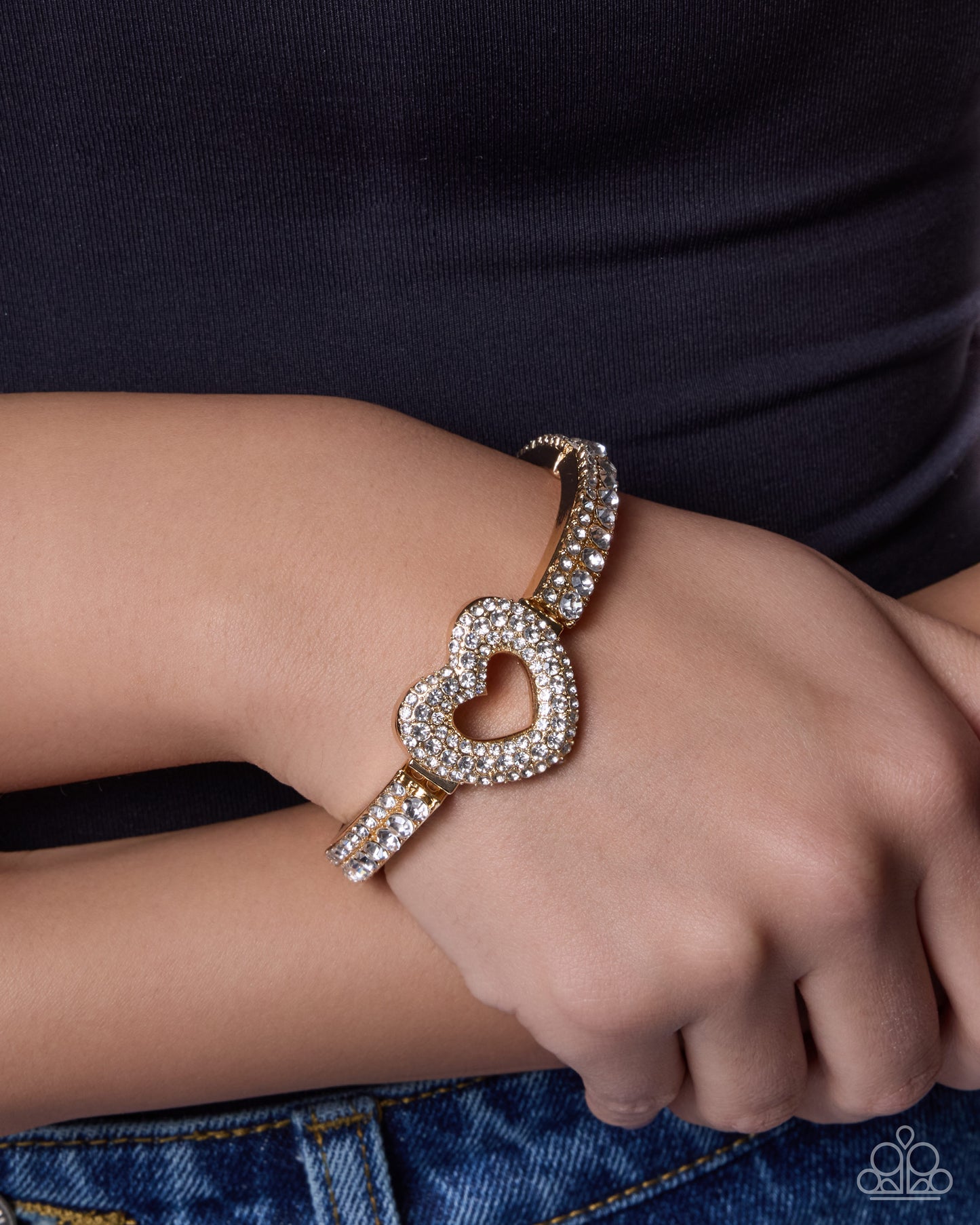 A close-up of a person's wrist wearing a gold heart-shaped cuff bracelet adorned with sparkling clear crystals, paired with a black top and blue jeans.