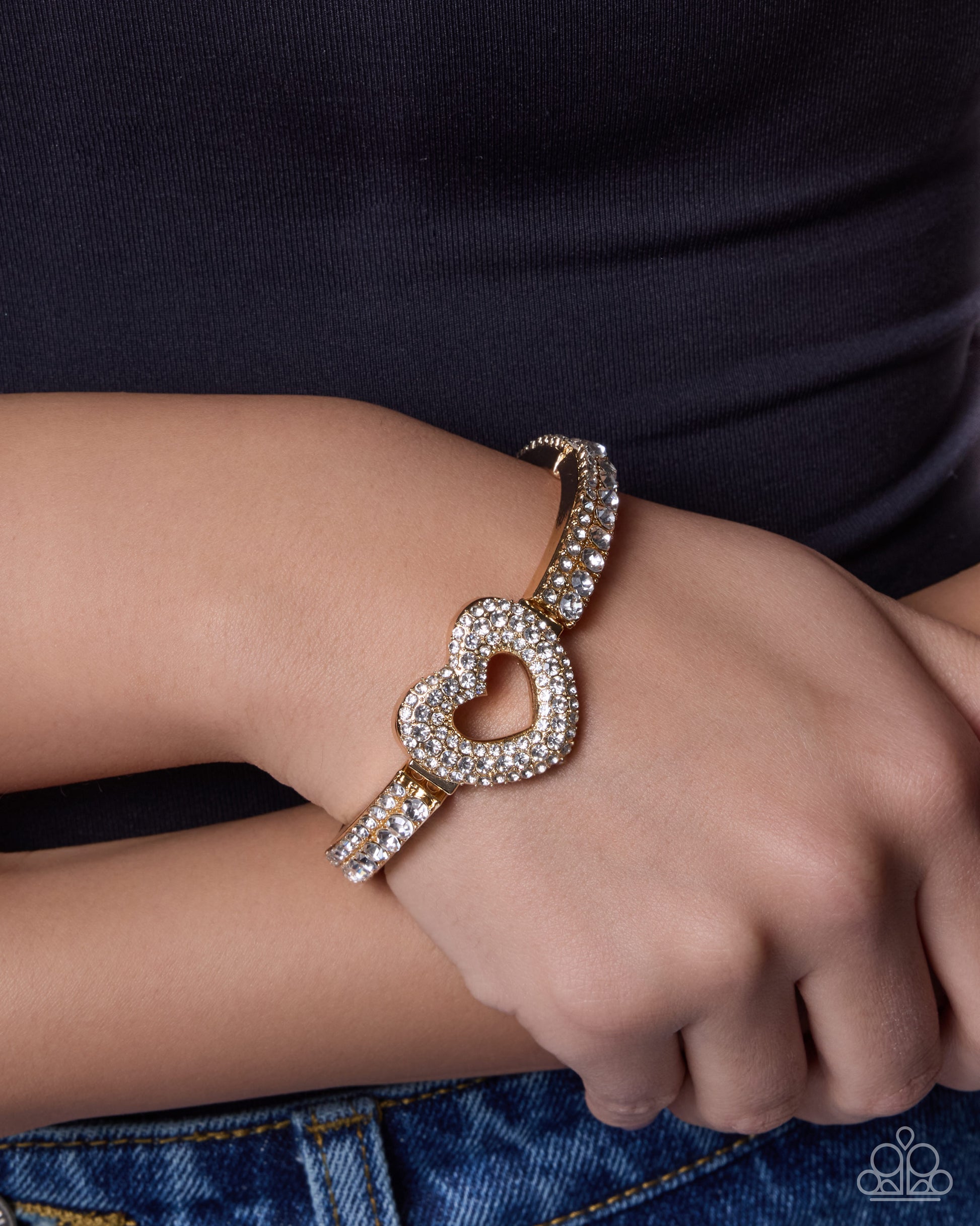 A close-up of a person's wrist wearing a gold heart-shaped cuff bracelet adorned with sparkling clear crystals, paired with a black top and blue jeans.