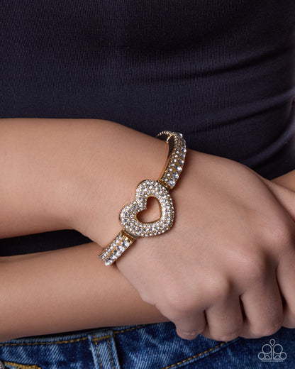 A close-up of a person's wrist wearing a gold heart-shaped cuff bracelet adorned with sparkling clear crystals, paired with a black top and blue jeans.