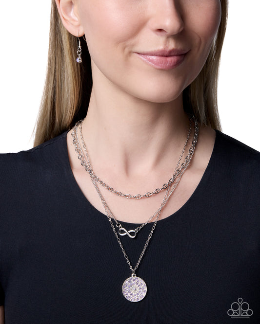 A close-up of a woman wearing a layered silver necklace set with a round pavé disc pendant, an infinity link, and a delicate chain, along with a matching drop earring, against a white background.