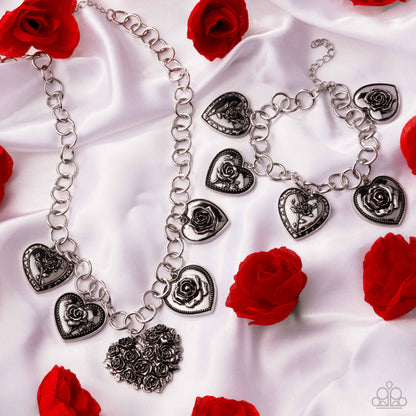 Silver necklace with heart and rose charms on a white fabric background with red roses.