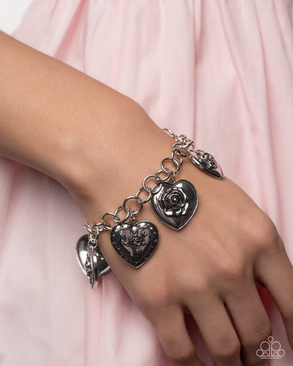 Silver heart-shaped charm bracelet on a wrist with a pink fabric background