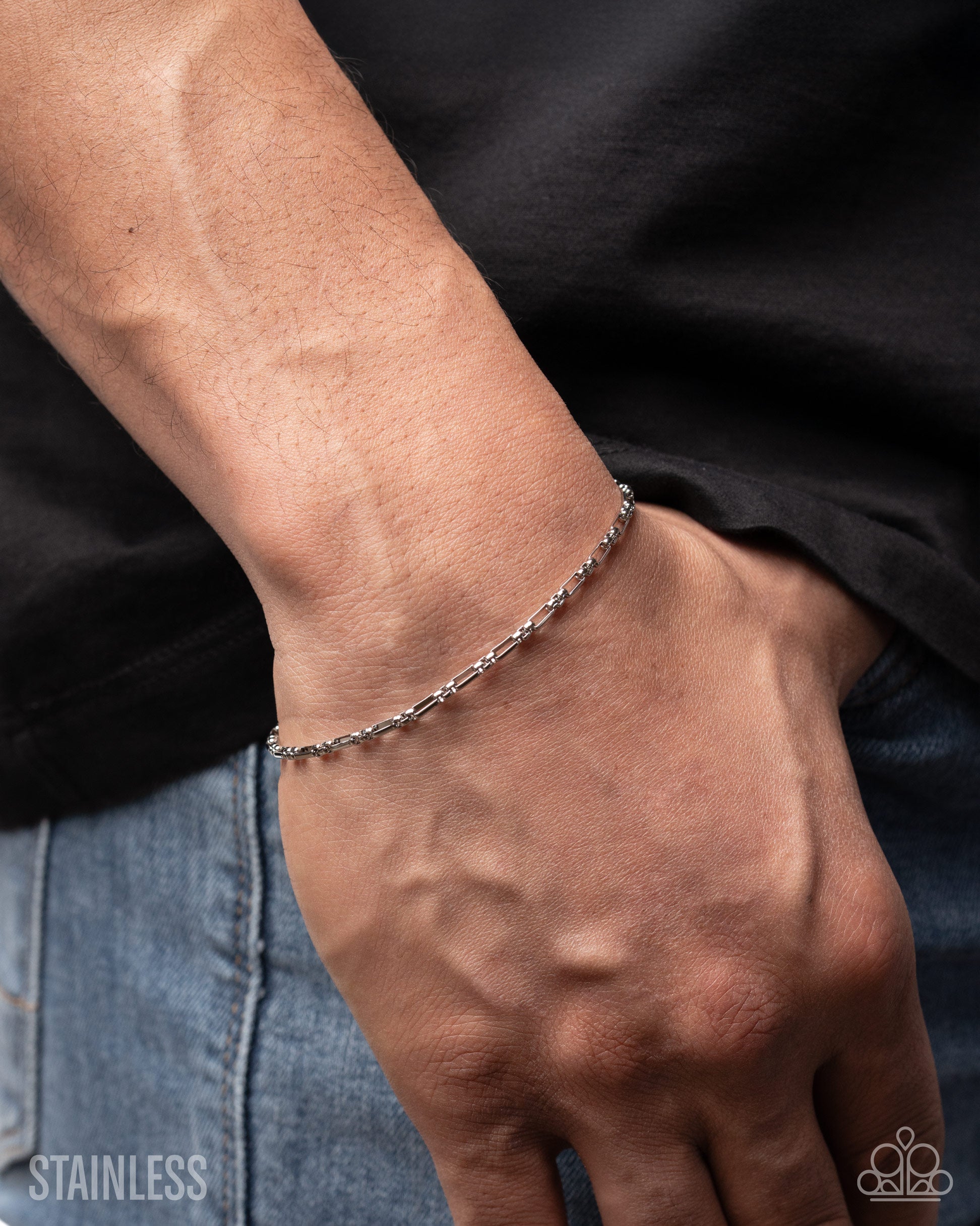 Close-up of a person's wrist wearing a silver chain bracelet with a black shirt and blue jeans in the background.