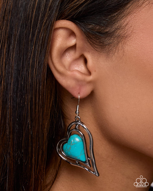 A close-up of a model's ear wearing a silver heart-shaped dangle earring with a turquoise center stone. The image features a warm, clean background with the model's dark brown hair framing the side profile.