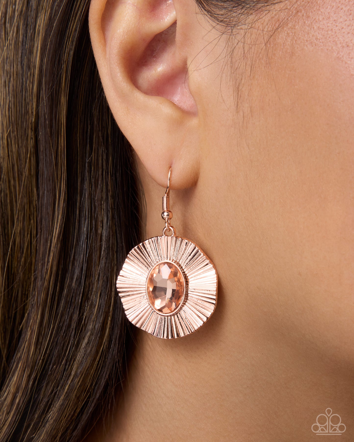A close-up of a model's ear wearing a single rose gold dangle earring with a sunburst disc design and a peach-colored oval gemstone.